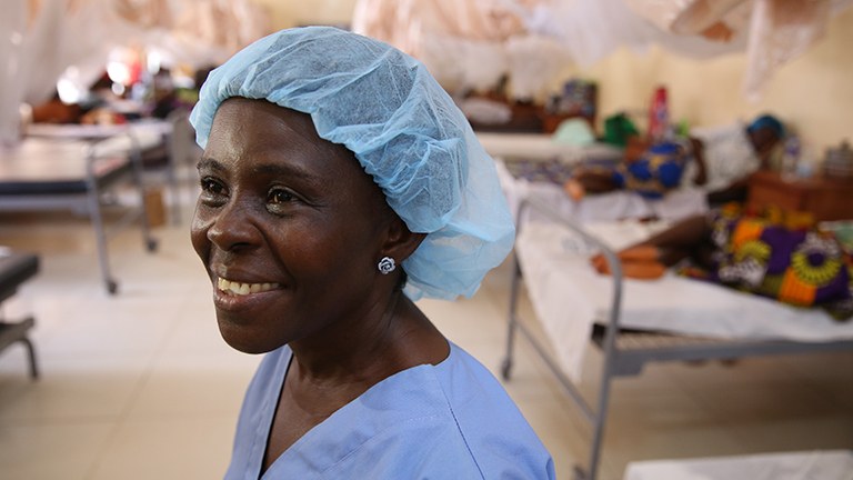 Image of a female healthcare worker in a hospital with women in beds behind her. 