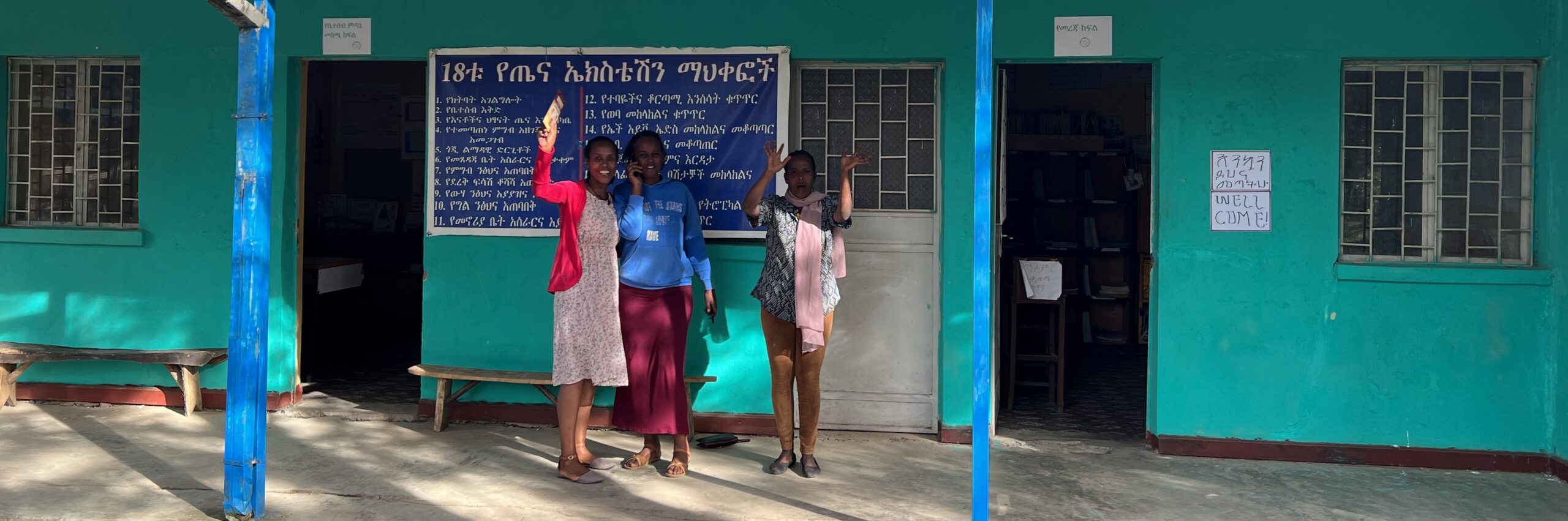 Photo of three women smiling and waving from the porch of a small health facility in Ethiopia.