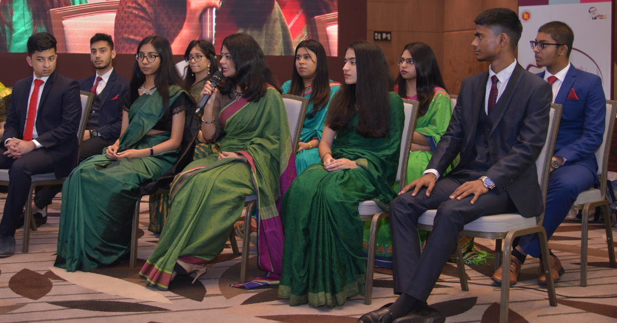 Photo showing a group of adolescents sitting in an auditorium, with one of them speaking into a handheld microphone, while her image is projected to a screen nearby.