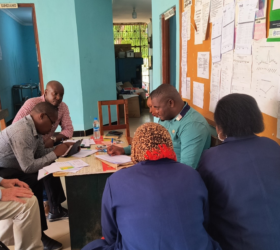 Photo showing a group of six Tanzanian researchers seated around a wooden table taking notes.
