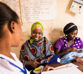 Photo showing two moms in a clinic in Tanzania, one smiling at a partially visible health care worker and one gazing at her infant.