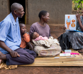 Photo showing a Ugandan health care worker talking to a family consisting of a man, woman, young child, and infant.